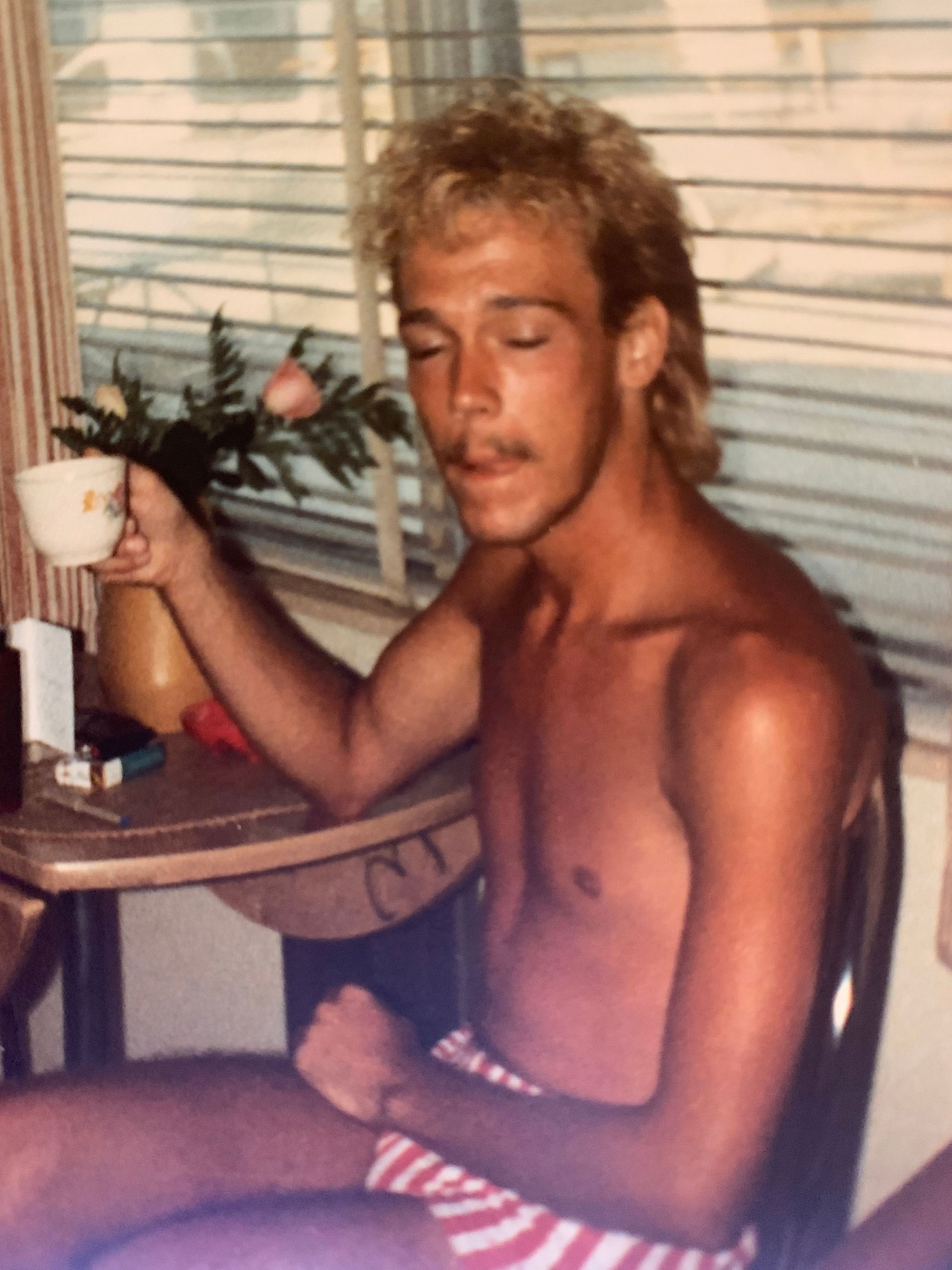 a very tan white man with bleached blonde hair is sitting in a chair next to a table sipping tea. he is wearing a small pair of white and red striped shorts.