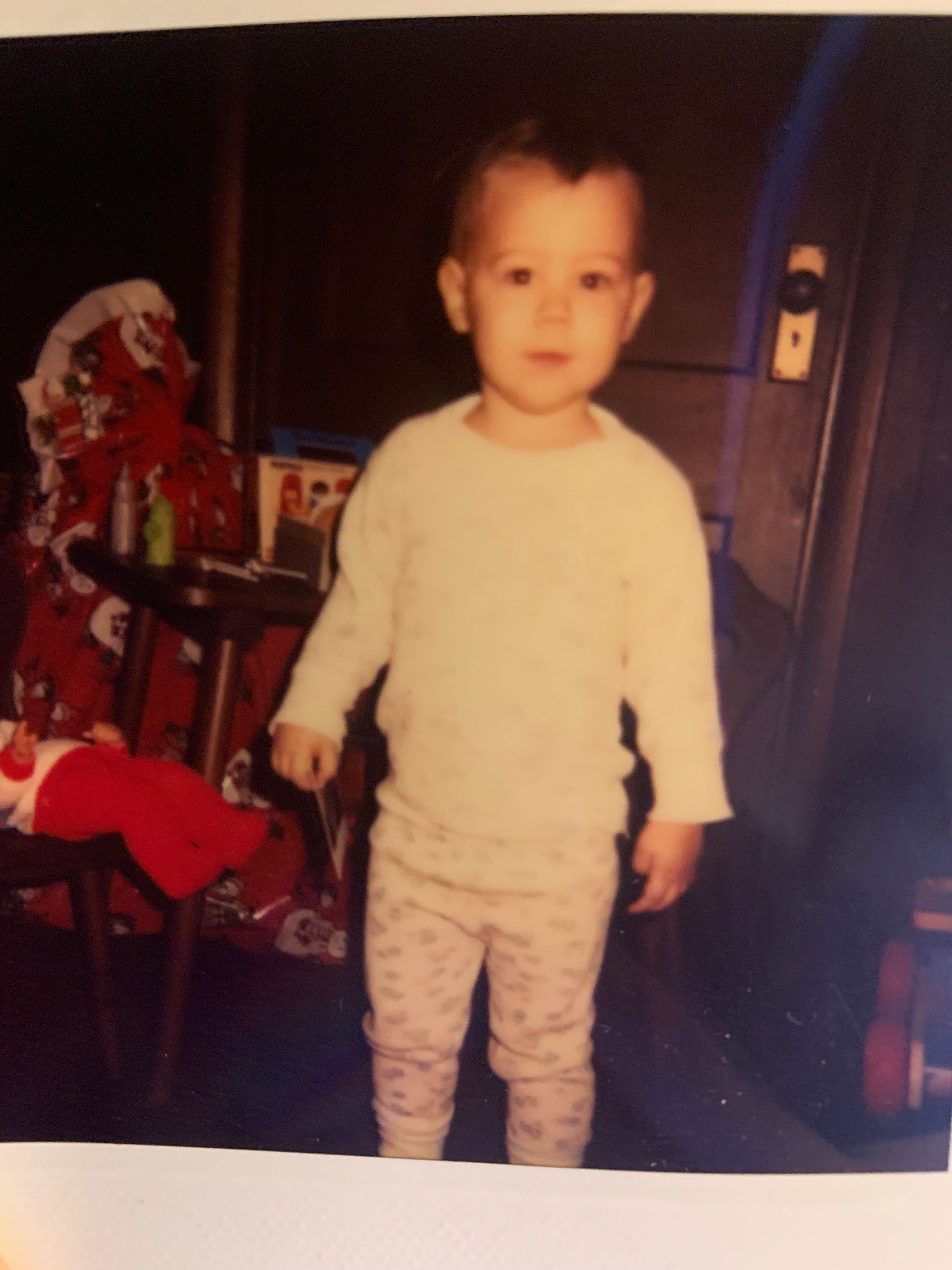 a white baby, nearly 1 year old, wearing a white shirt and patterned pants, standing in front of some xmas decorations