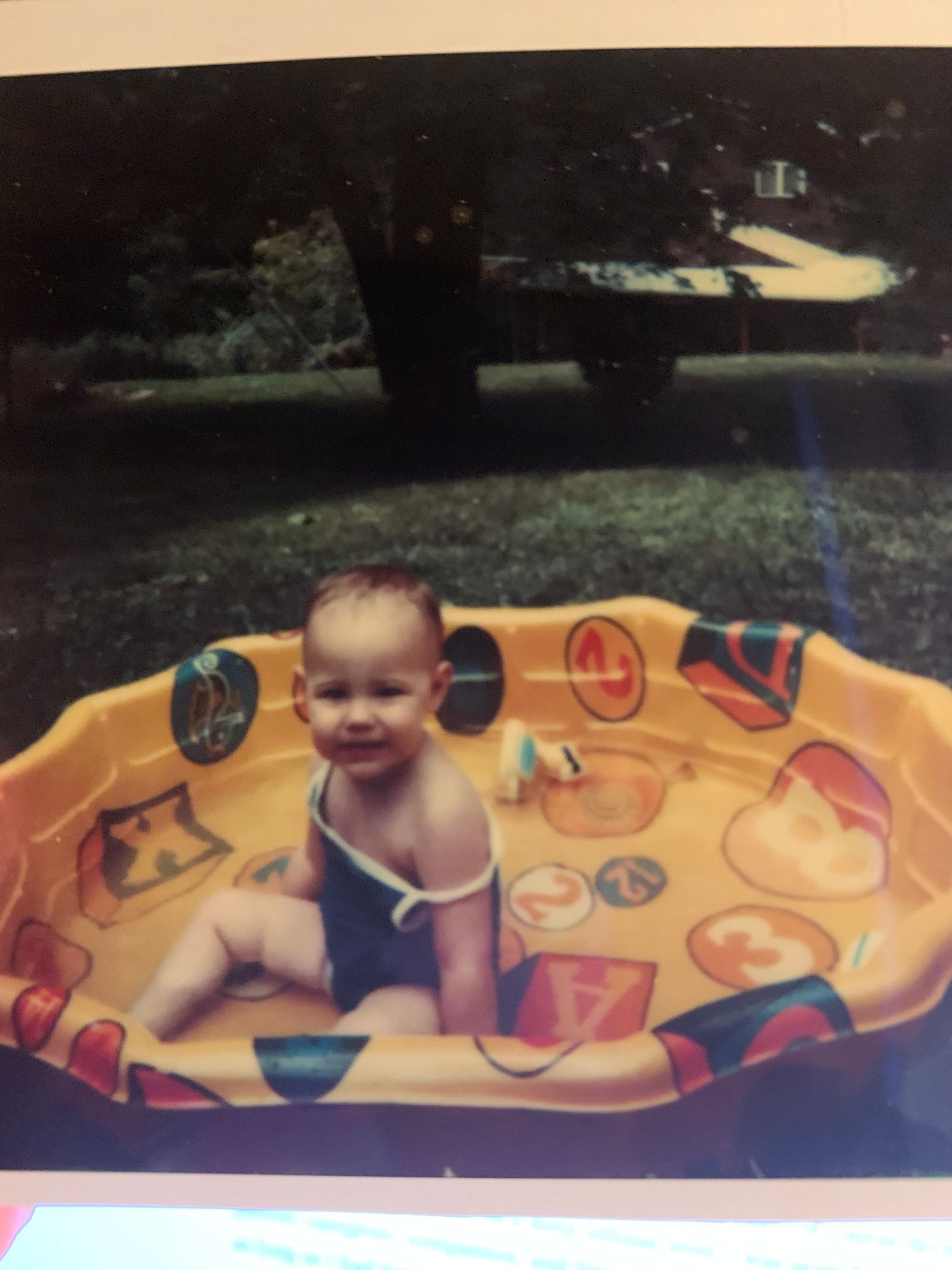 a chubby white baby with barely-there dark brown hair wearing a blue and white bathing suit, sitting in a yellow plastic pool with a few inches of water and some toys in it.