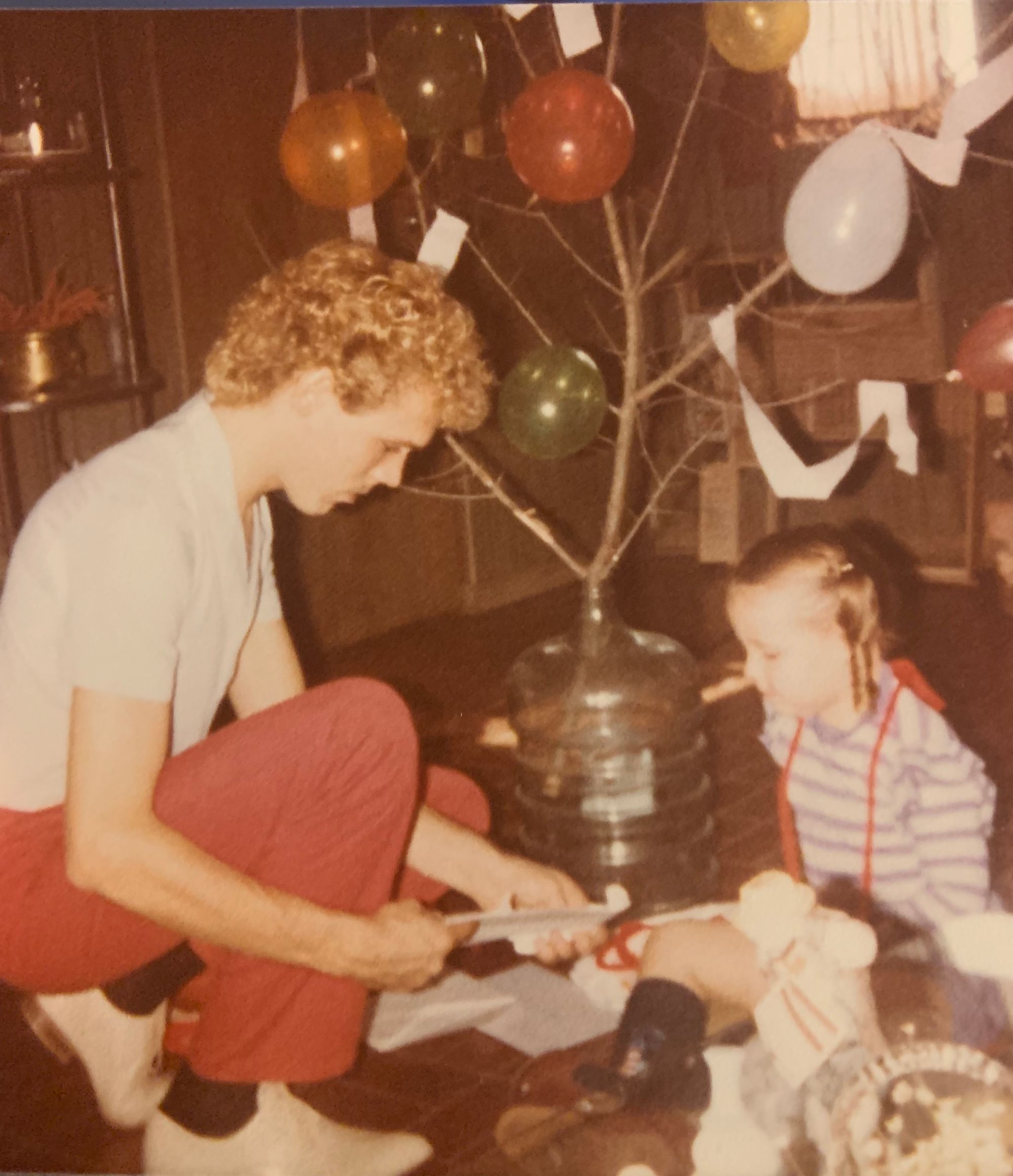 a thin white man with bleached blonde hair kneels and shows something to a little kid sitting opposite him. the little kid is wearing a purple striped shirt. in between them is a tree branch with multicolored balloons attached all over it.