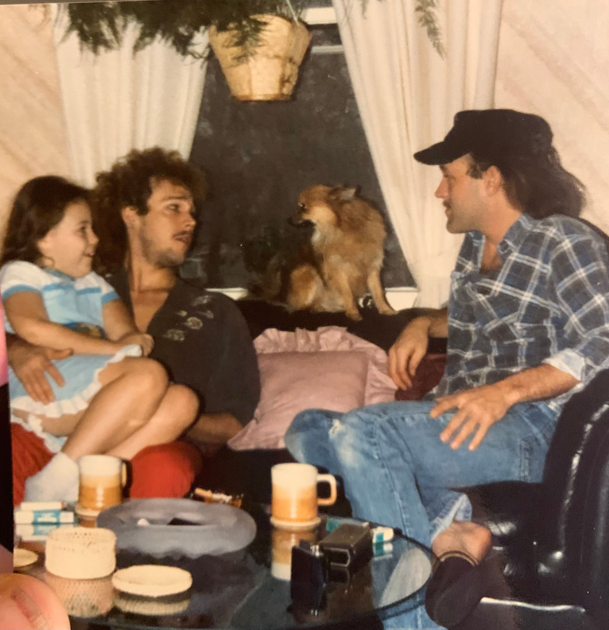 a white kid with long dark brown hair is wearing a blue and white nightgown and is sitting on the lap of a white man wearing a black button-down shirt. there is a dog looking at both of them. another white man, this one with longer brown hair, wears a gray flannel shirt and jeans, and is also focused on the dog.