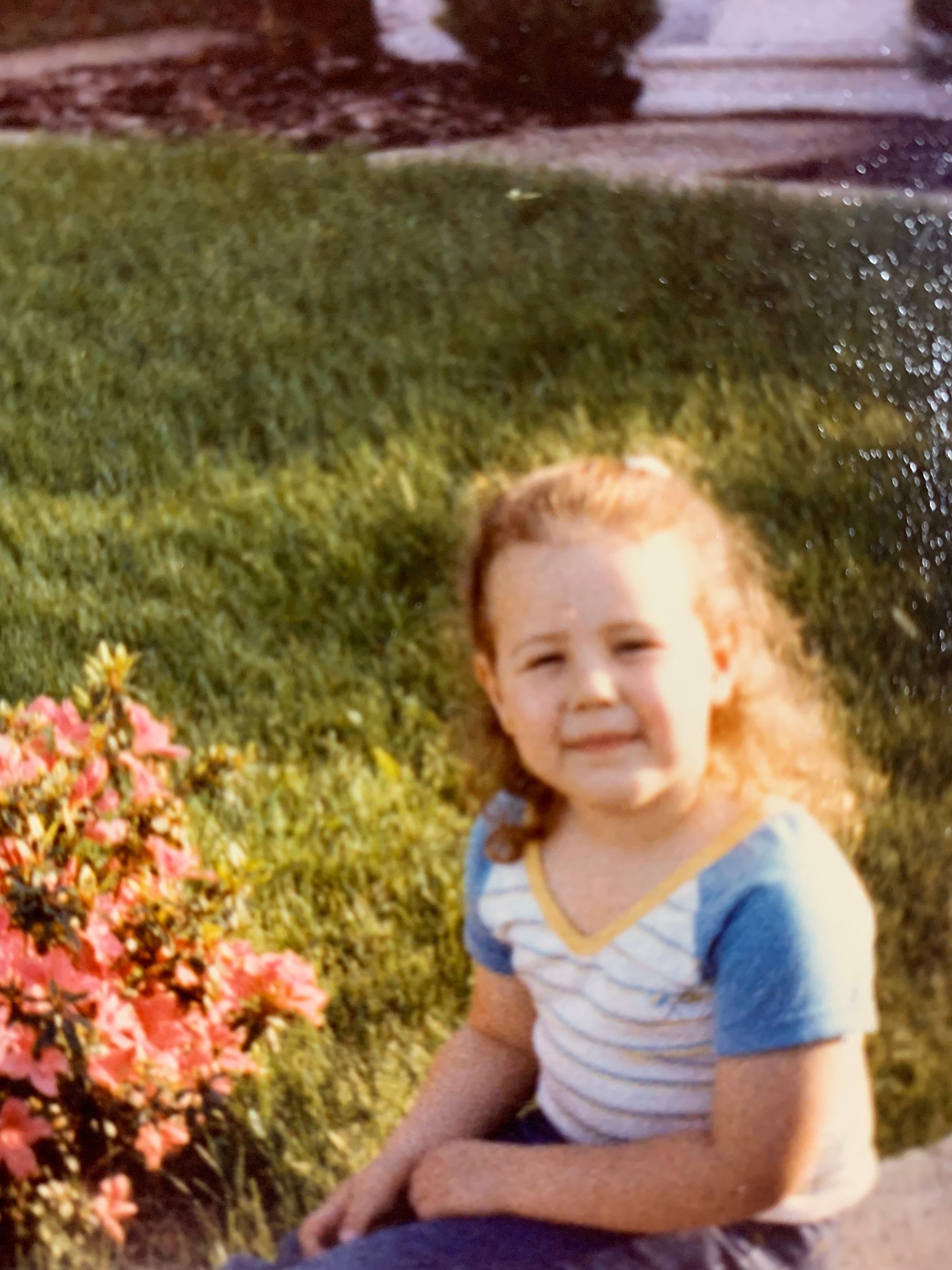 a white brown haired child sits near a pink flowering bush, wearing a blue yellow and white shit and blue pants. zi is smiling in the picture.