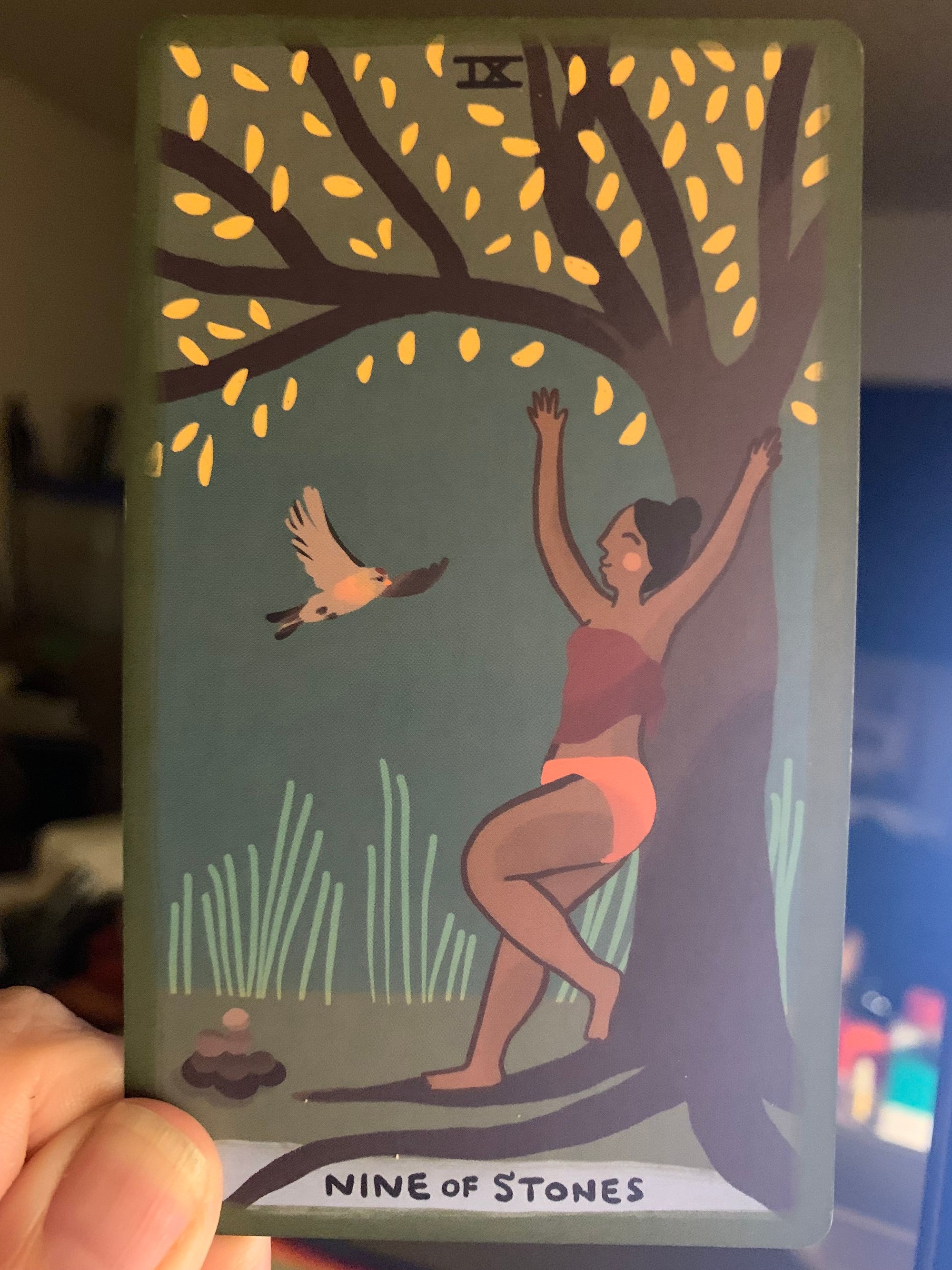 A tarot card, the nine of stones, featuring a brown smiling person beneath a tree with their arms up. Tall grasses grow behind the person and their pile of nine stones, and the tree hangs glorious golden leaves above them.